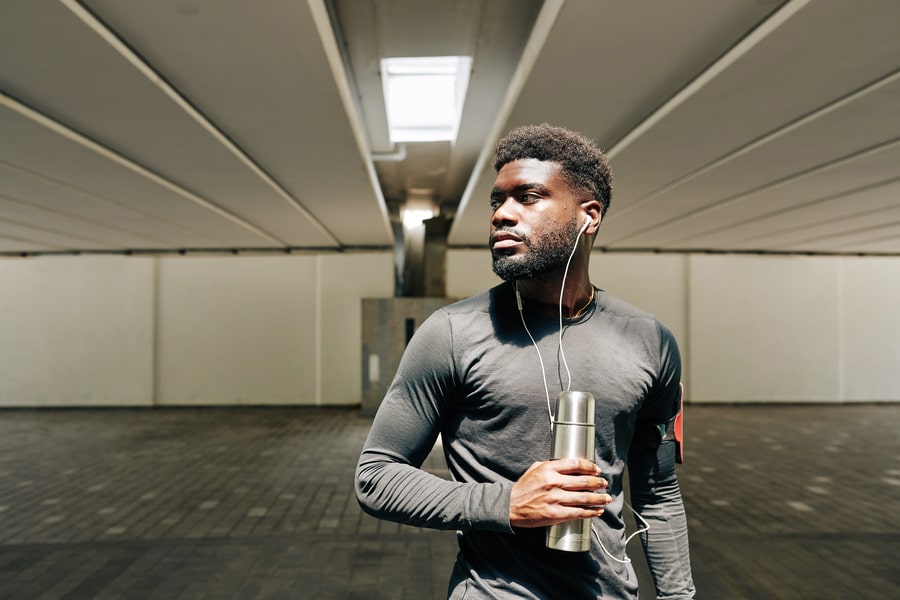 Portrait of serious young Black sportsman listening to music with earbuds and drinking water.