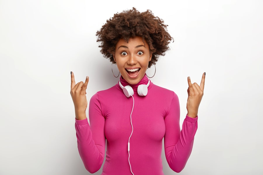 Pretty Afro American girl with curly hair, makes rock n roll gesture, enjoys listening favourite music.